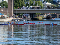 Kanufahrerinnen und -fahrer auf einem Fluss beim Ufer, im Hintergrund eine Brücke.