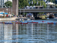 Kanufahrerinnen und -fahrer auf einem Fluss beim Ufer, im Hintergrund eine Brücke.