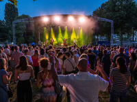 Auf einer gelb erleuchteten Bühne im Hintergrund spielt eine Band, davor steht Publikum mit teilweise tanzenden Menschen.
