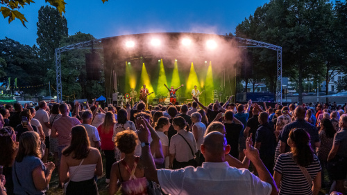 Auf einer gelb erleuchteten Bühne im Hintergrund spielt eine Band, davor steht Publikum mit teilweise tanzenden Menschen.