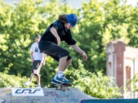 Skateboarder mit blauem Hut vollführt einen Trick über eine Rampe im Skatepark vor grünen Bäumen und Graffiti.
