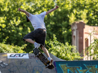 Skateboarder springt im Skatepark über eine Rampe, umgeben von Graffiti und Bäumen im Hintergrund.