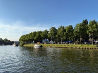 Ein Boot auf einem Fluss, daneben Stände am Ufer. Dahinter Bäume und der Himmel.