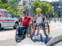 Ein Mädchen mit Helm und Rollstuhl fährt über eine Rampe, neben ihr fährt ein Mann mit Rollstuhl.