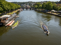 Zwei Drachenboote fahren auf einem Fluss, links am Ufer sind Boote, rechts am Ufer verschiedene Stände.