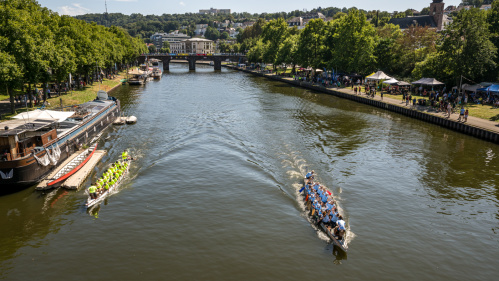 Zwei Drachenboote fahren auf einem Fluss, links am Ufer sind Boote, rechts am Ufer verschiedene Stände.