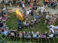 Draufsicht von oben auf eine Wiese mit einem Halbkreis aus Klappstühlen, auf denen Menschen sitzen. Dahinter sitzen Personen an Biergarnituren.