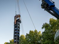 Ein Kind steht auf einem Turm aus Getränkekisten, zwei Männer in Uniformen sichern es mit Seilen an einem Kran. Dahinter stehen weitere Personen.