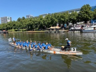 Ein Drachenboot fährt auf einem Fluss, am Ufer dahinter sind Stände und auf dem Wasser am FlussrandBoote.