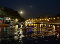 Drachenboote fahren im Dunklen auf einem Fluss, rechts davon stehen Boote. Links sind gelbe Lichter im Hintergrund und am Himmel der Vollmond, auf dem Wasser ist gelber Lichterglanz.