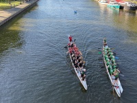 Zwei Drachenboote fahren auf einem Fluss, links und rechts im Hintergrund sind Bäume und Stände am Ufer , rechts sind am Rand des Flusses Boote.