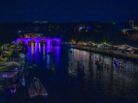 Drachenboote fahren nachts auf einem Fluss, dahinter eine beleuchtete Brücke. Am Ufer sind Boote und auf dem Uferbereich sind beleuchtete Stände.