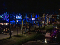 Viele Menschen und Stände nachts am Flussufer mit Lichterketten und beleuchteten Bäumen. Rechts davon auf dem Wasser sind Boote.