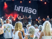 Eine Band spielt auf einer Bühne, davor steht Publikum. Auf einem Banner auf der Bühne steht das Wort bruise.