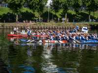  Drachenboote fahren nebeneinander auf einem Fluss. Dahinter Böume am Ufer.