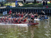 Menschen in Drachenbooten rudeln auf einem Fluss, dahinter am Ufer sitzen und stehen Zuschauende.