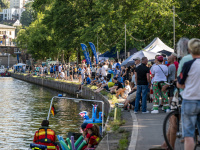 Zahlreiche Menschen stehen an einem Flussufer, dahinter Bäume und eine Brücke.