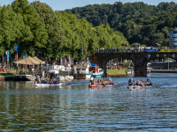 Drei Drachenboote auf einem Fluss, am Ufer liegen Boote auf dem Wasser. Dahinter Bäume und eine Brücke.