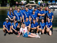 Menschen in blauen Trickots posieren in mehreren Reihen für ein Teamfoto vor dem Wasser.