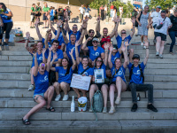 Menschen in blauen Trickots posieren in mehreren Reihen für ein Teamfoto auf einer Treppe.